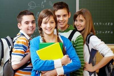 Group of smiling school-aged children standing in front of a chalkboard with backpacks and notebooks