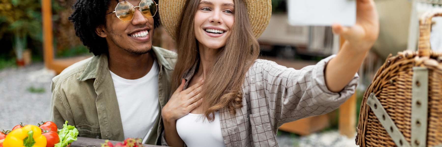 Smiling young couple taking a selfie outdoors at a picnic table with fresh vegetables and a wicker b